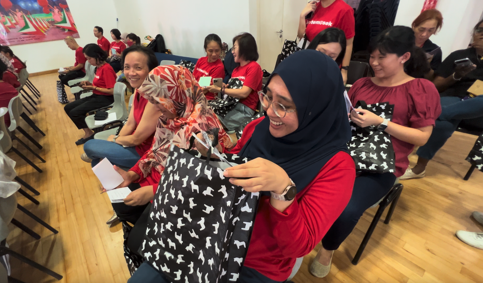 A smiling woman in a navy hijab holds a black patterned bag at a gathering with others in red shirts.